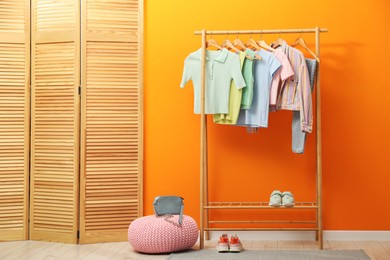 Rack with different stylish women's clothes, shoes, bag and pouf near orange wall indoors