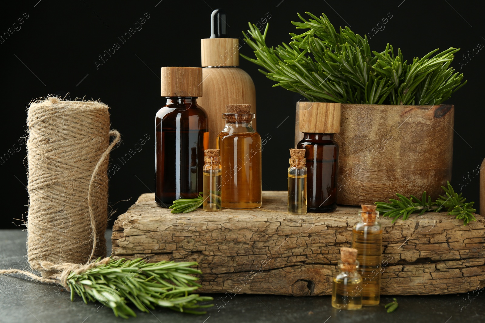 Photo of Essential oils in bottles, rosemary, thread and wood on gray table against black background