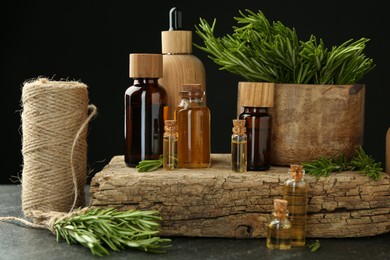 Photo of Essential oils in bottles, rosemary, thread and wood on gray table against black background