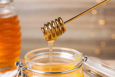 Photo of Dripping tasty honey from dipper into jar on blurred background, closeup