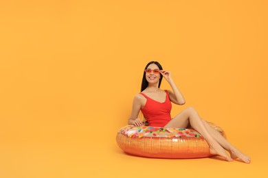 Photo of Happy young woman with beautiful suntan and sunglasses on inflatable ring against orange background, space for text