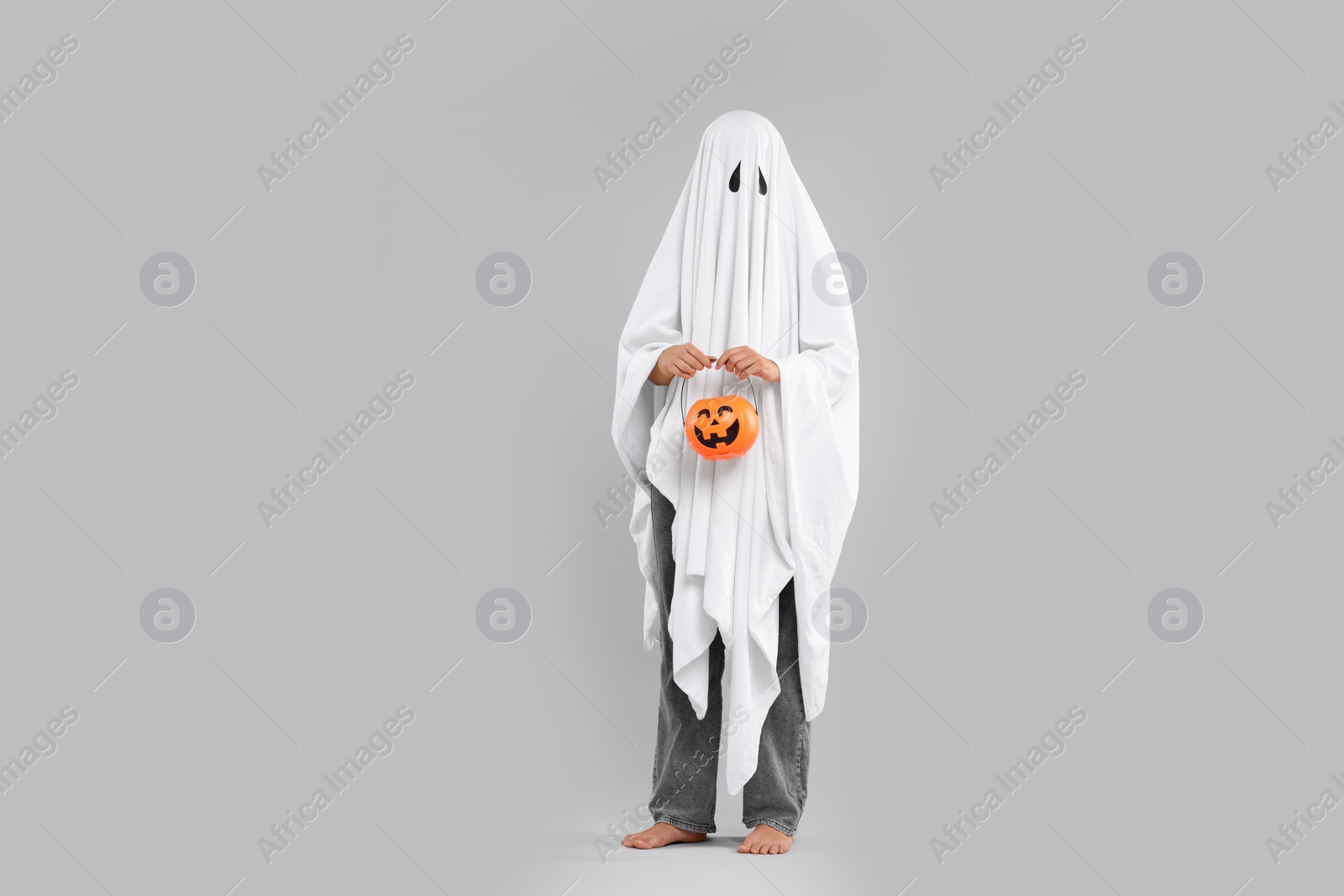 Photo of Woman in white ghost costume holding pumpkin bucket on light grey background. Halloween celebration