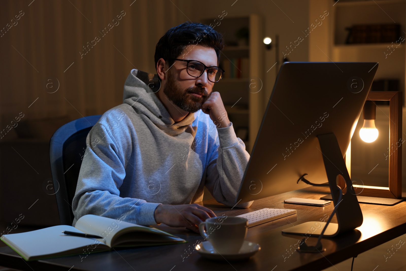 Photo of Home workplace. Tired man working with computer at wooden desk in room at night