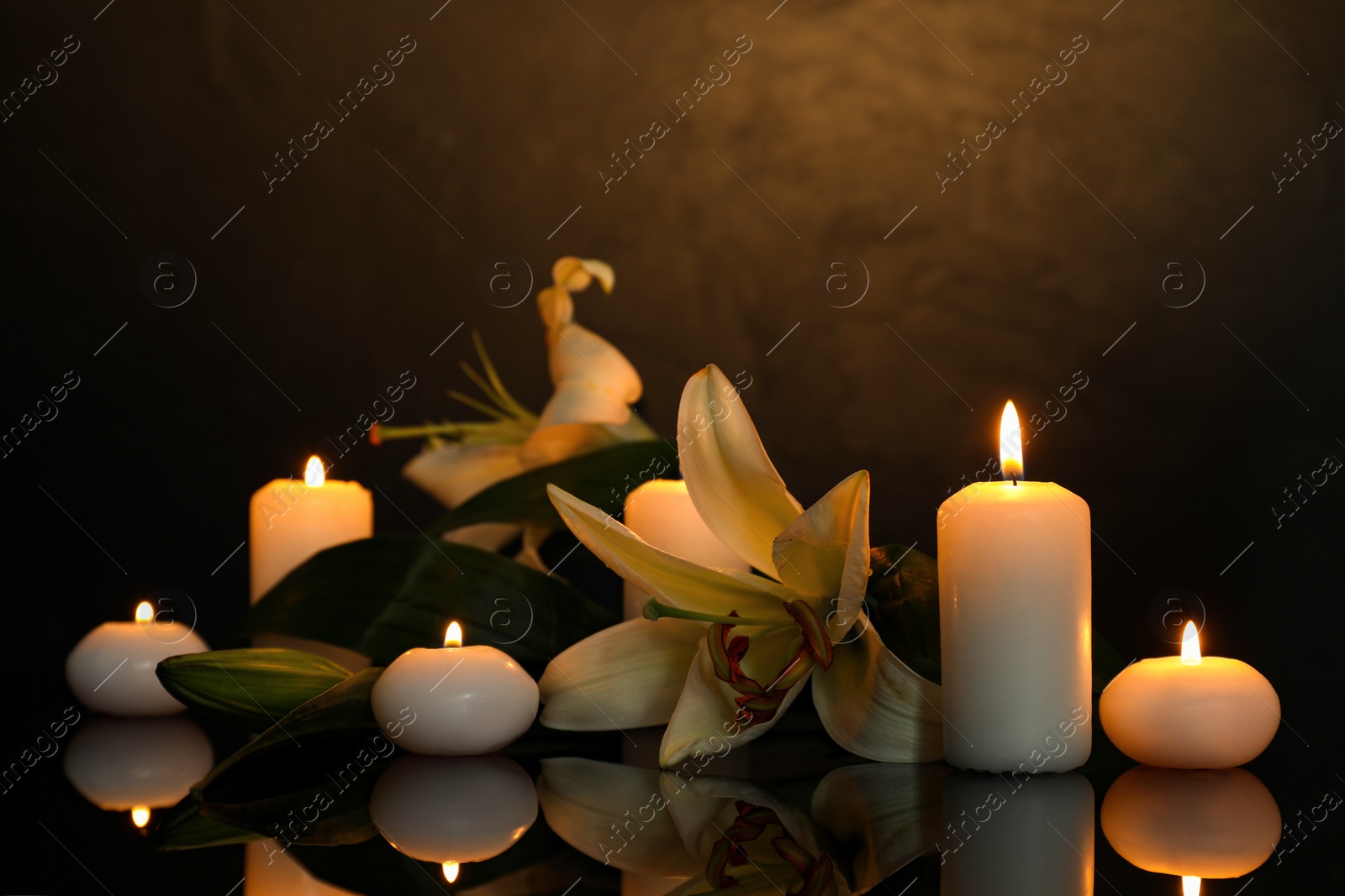Photo of White lilies and burning candles on black mirror surface in darkness, space for text. Funeral symbols