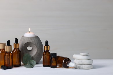 Photo of Different aromatherapy products, burning candle and eucalyptus leaves on white wooden table against light background, space for text