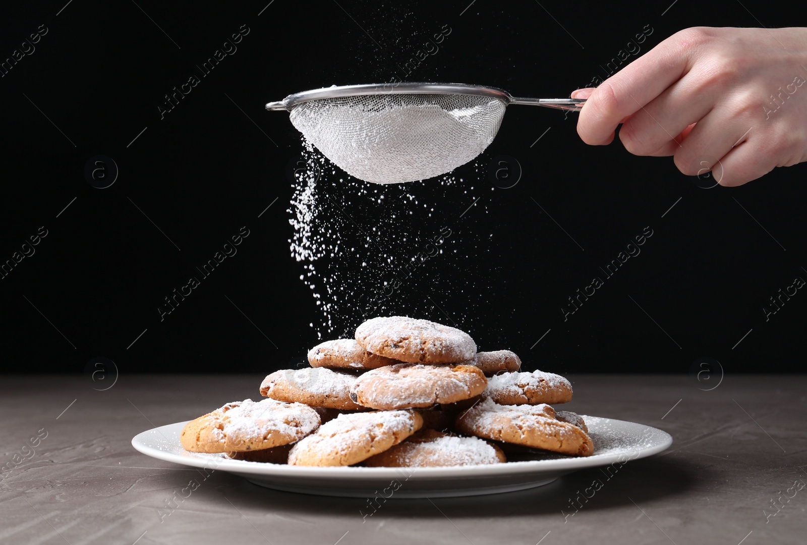 Photo of Woman with sieve sprinkling powdered sugar onto cookies at grey textured table, closeup