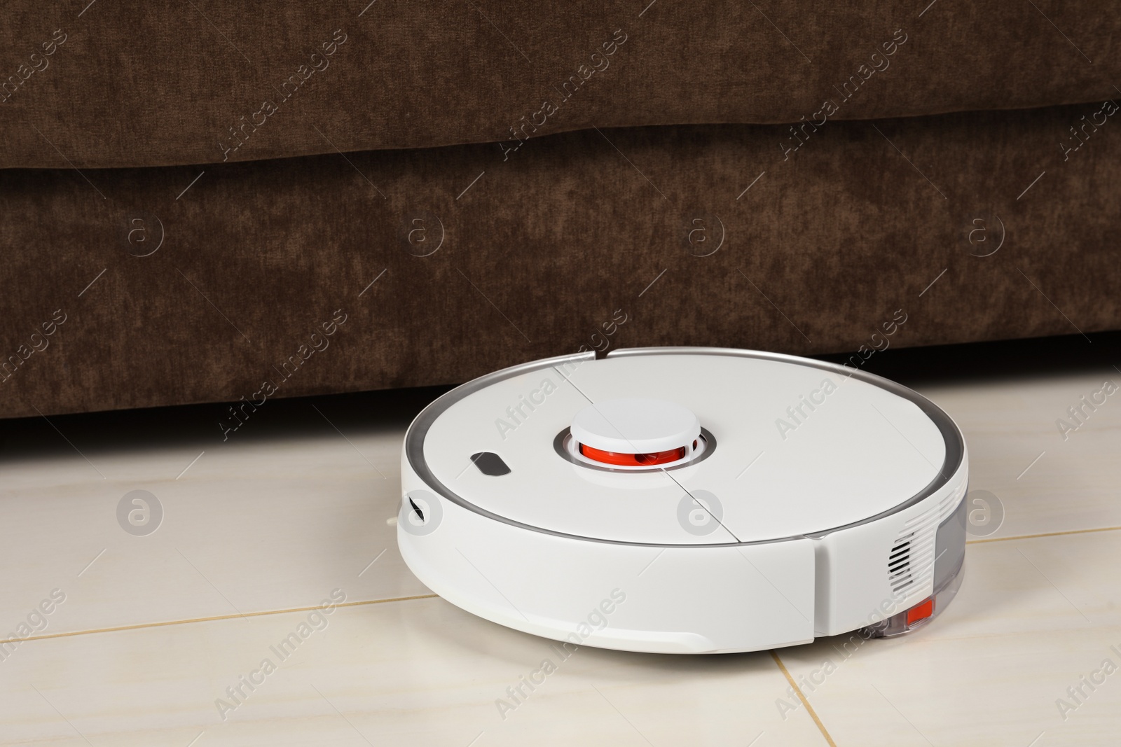 Photo of Robotic vacuum cleaner on white tiled floor near sofa indoors