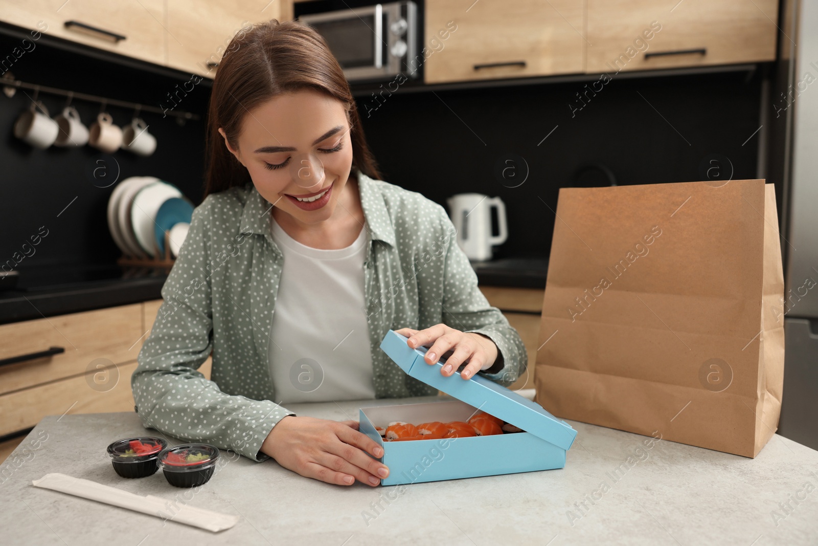 Photo of Beautiful young woman unpacking her order from sushi restaurant at table in kitchen