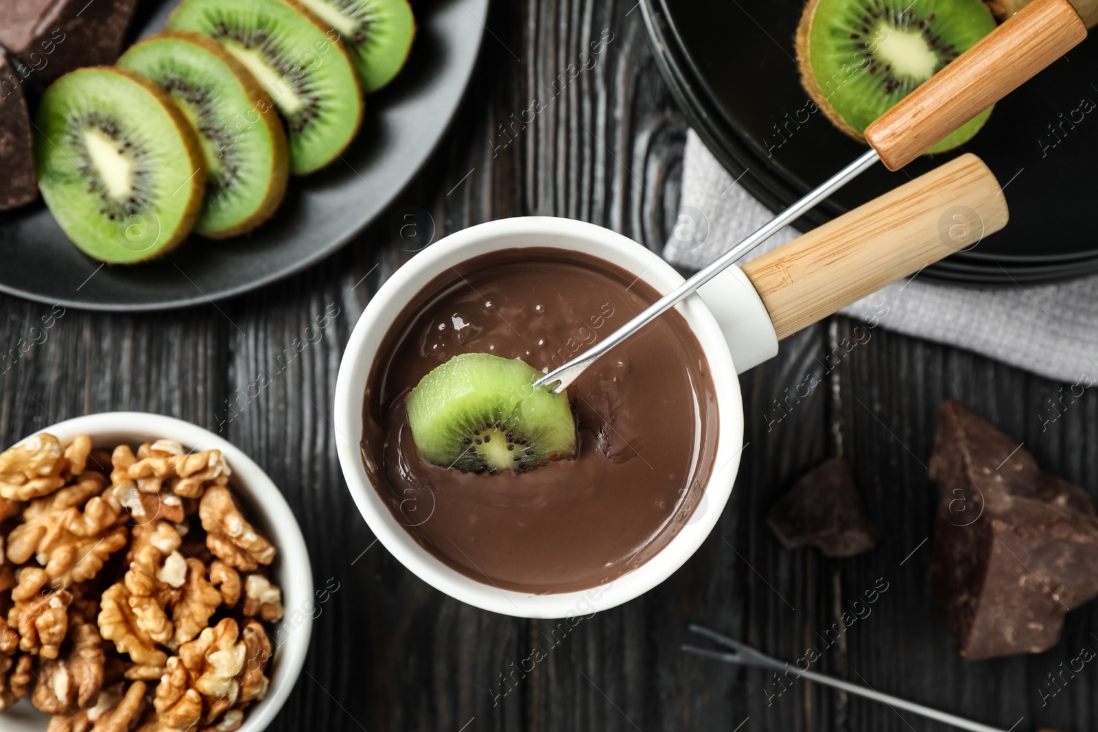 Photo of Dipping slice of kiwi into fondue pot with milk chocolate on black wooden table, top view