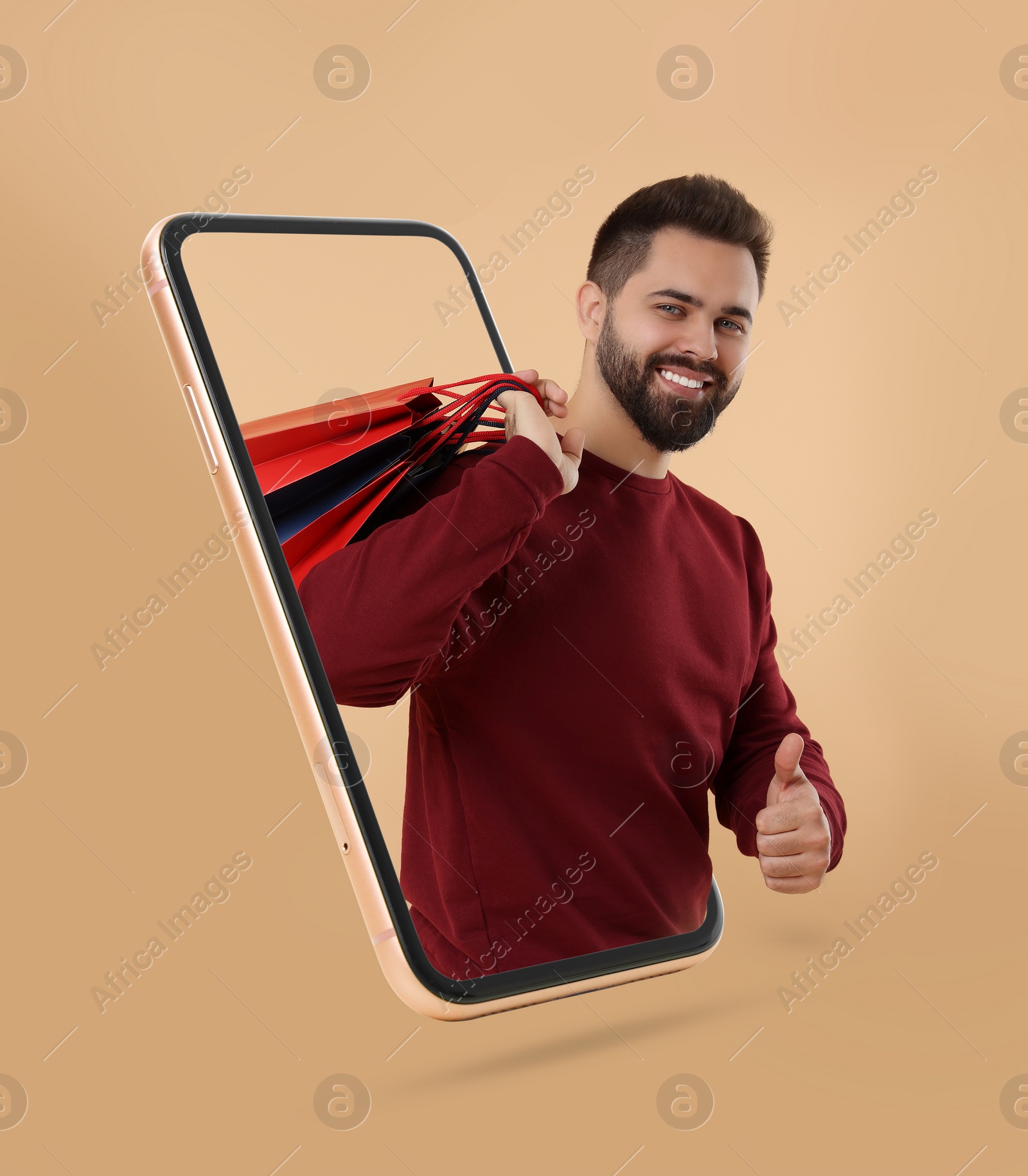 Image of Online shopping. Happy man with paper bags looking out from smartphone and showing thumbs up on beige background