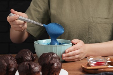 Photo of Woman mixing cream with blue food coloring at wooden table, closeup