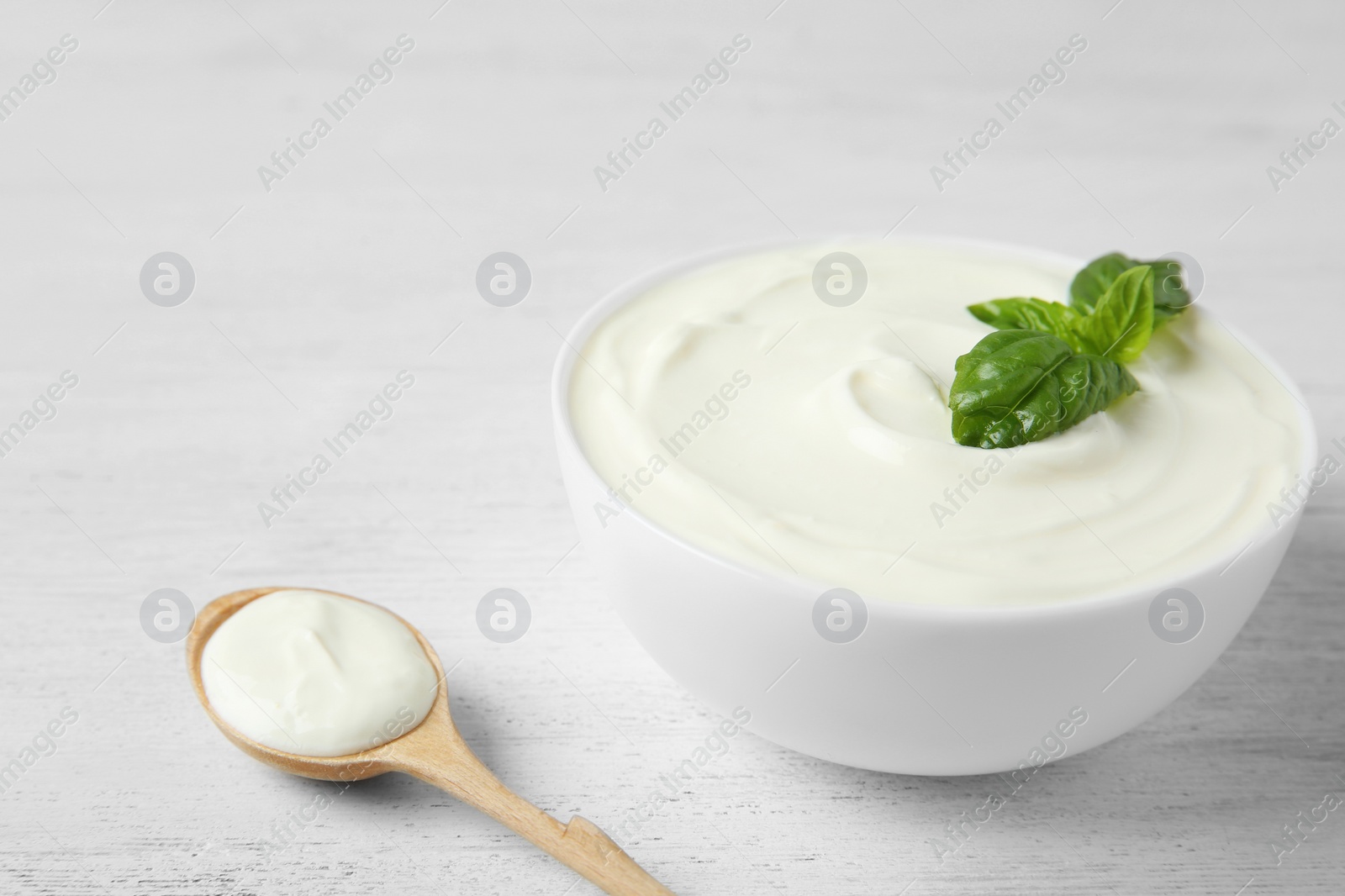 Photo of Bowl of fresh sour cream with basil and spoon on white wooden table. Space for text