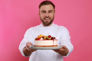 Photo of Happy professional confectioner in uniform holding delicious cake on pink background