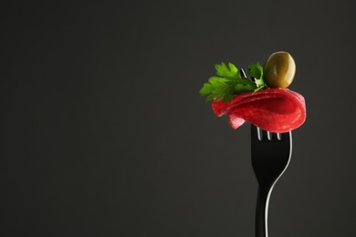 Photo of Fork with tasty slice of salami, olive and parsley on black background, space for text