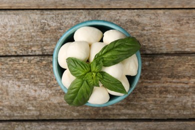 Photo of Tasty mozzarella balls and basil leaves in bowl on wooden table, top view