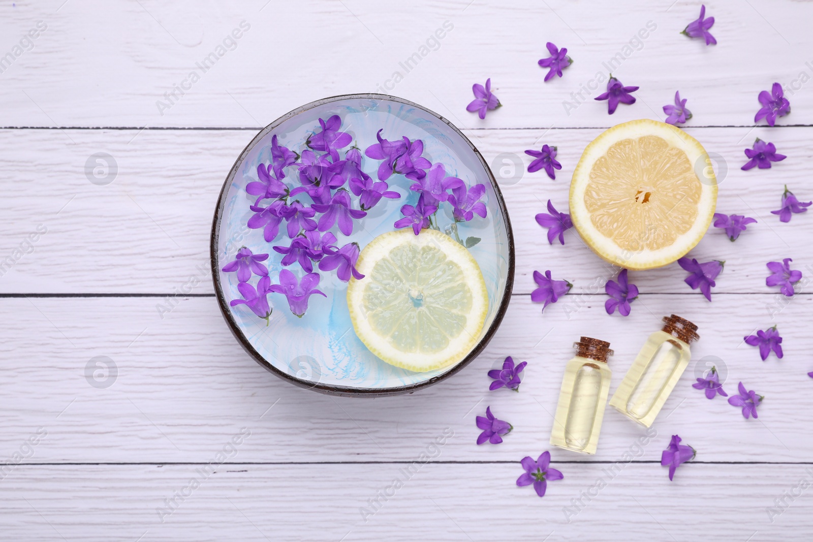 Photo of Bowl with water, lemon, purple flowers and bottles of essential oils on white wooden table, flat lay. Spa composition