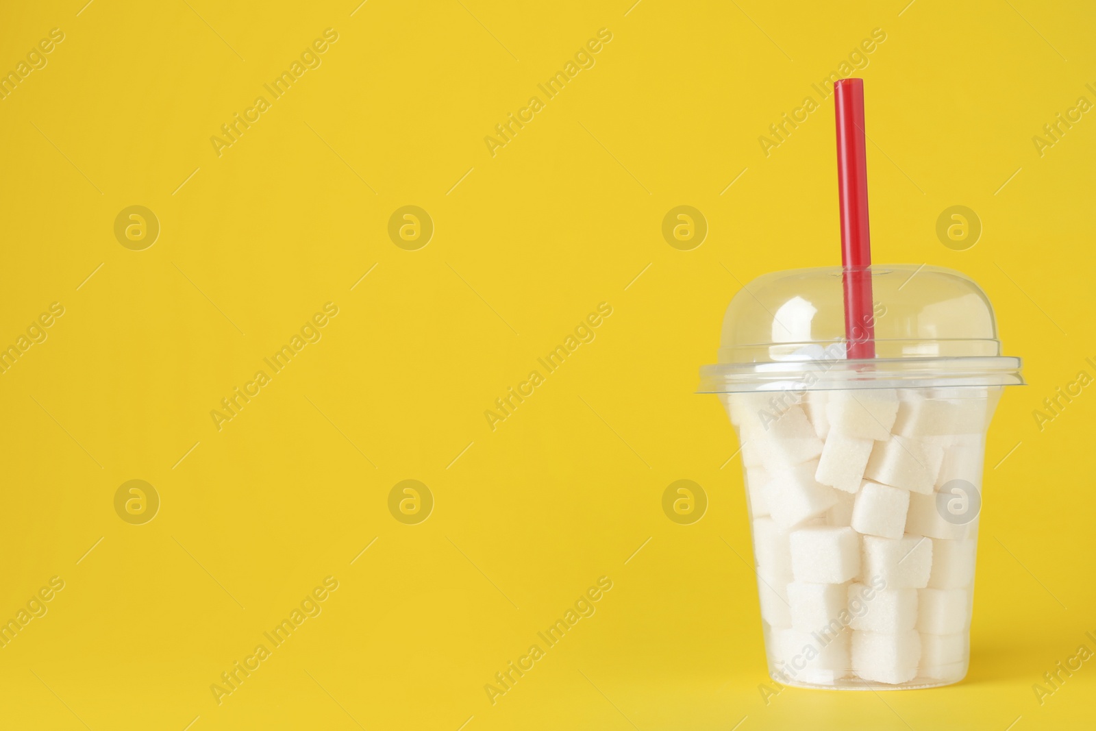 Photo of Refined sugar cubes in plastic cup with straw on yellow background. Space for text