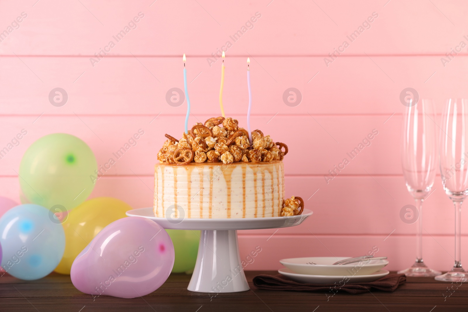 Photo of Caramel drip cake decorated with popcorn and pretzels near balloons and tableware on wooden table