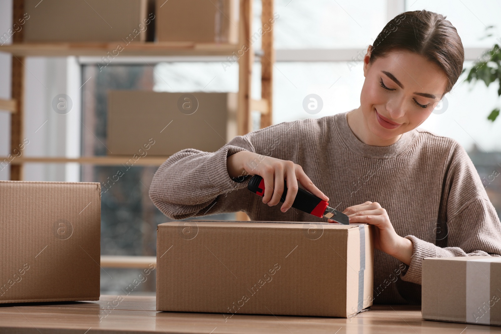 Photo of Young woman using utility knife to open parcel at wooden table indoors