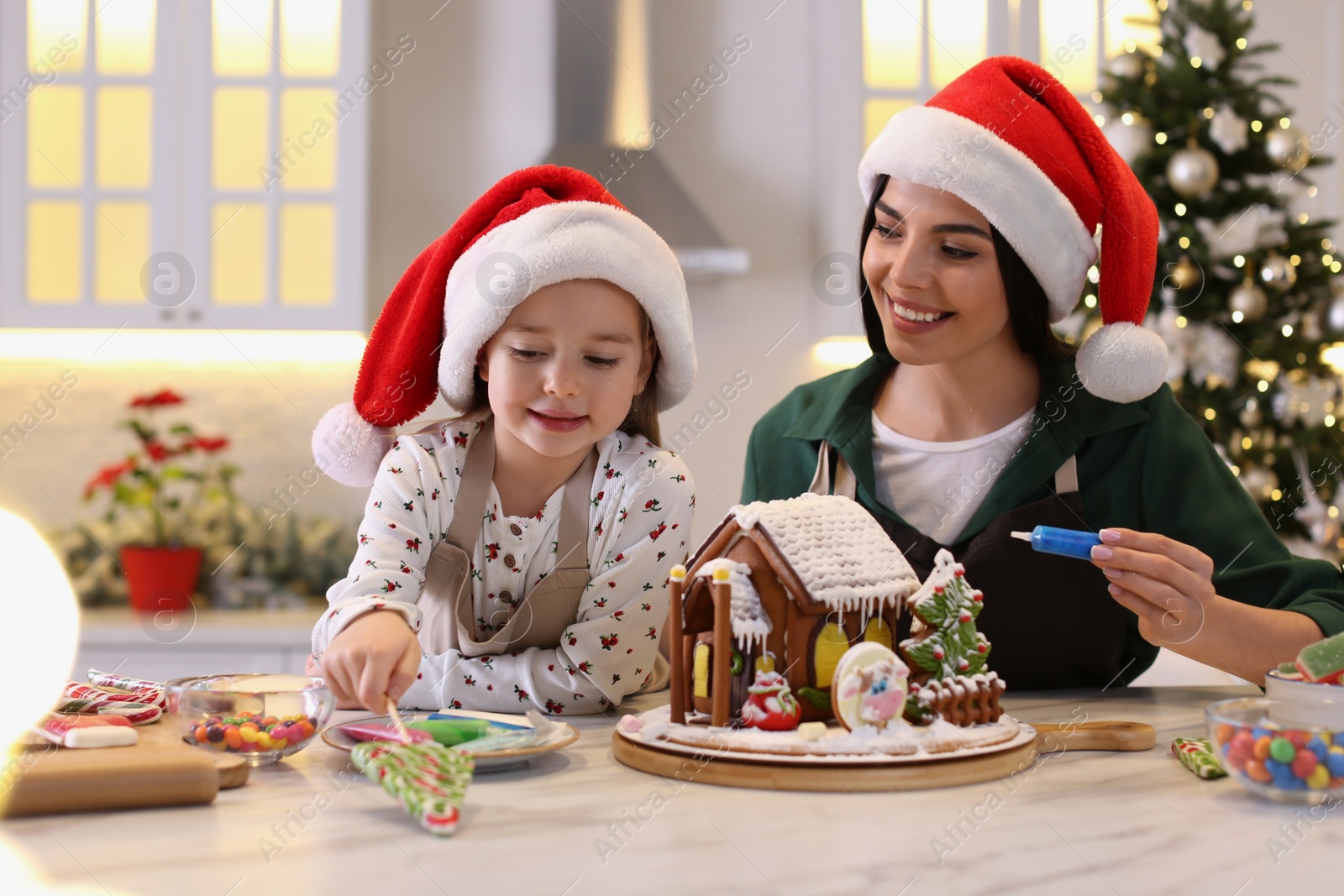 Photo of Mother and daughter decorating gingerbread house at table indoors