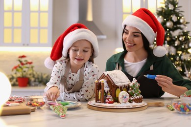 Photo of Mother and daughter decorating gingerbread house at table indoors