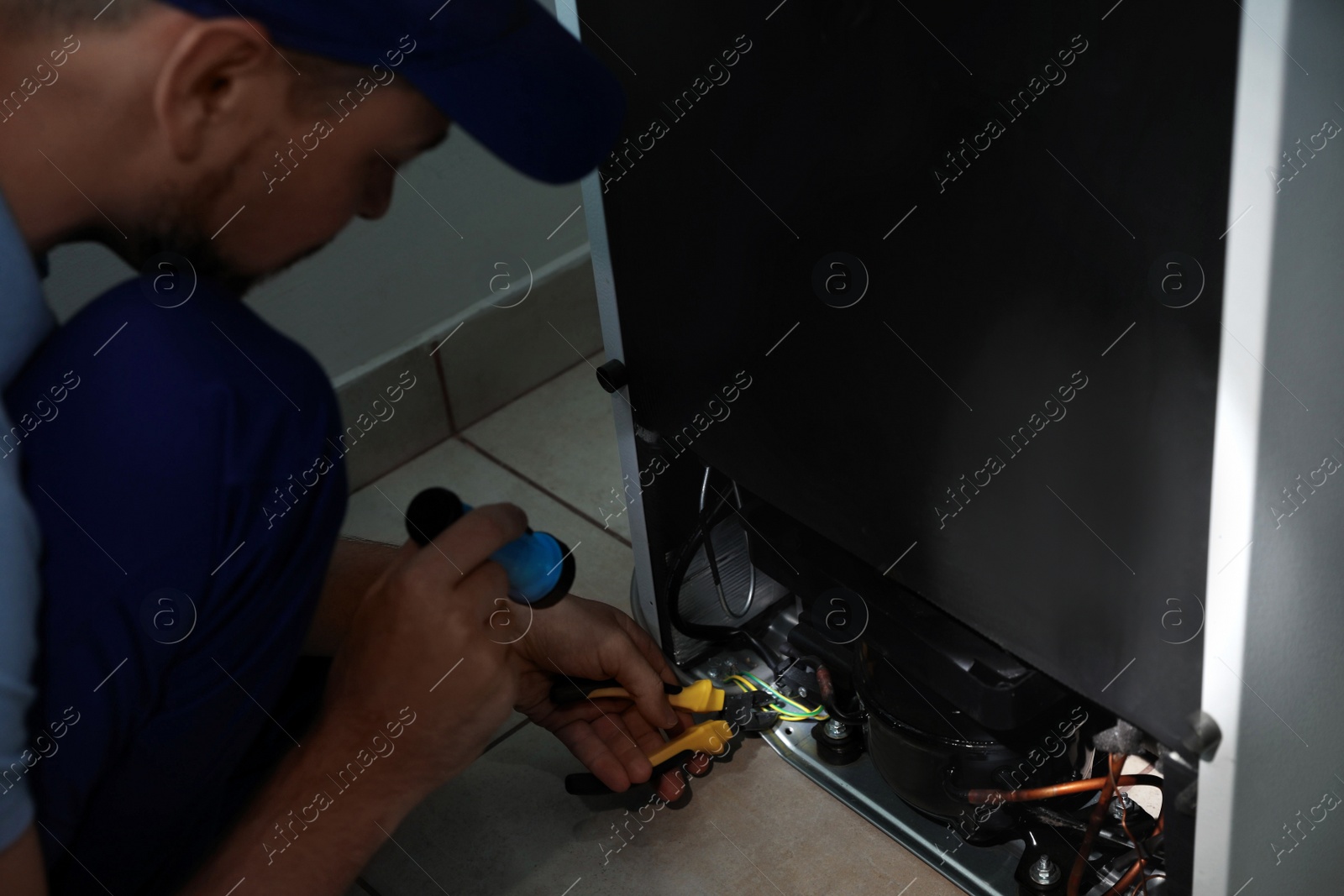 Photo of Electrician with flashlight fixing refrigerator indoors, closeup