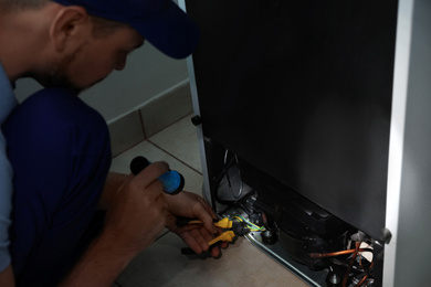 Photo of Electrician with flashlight fixing refrigerator indoors, closeup