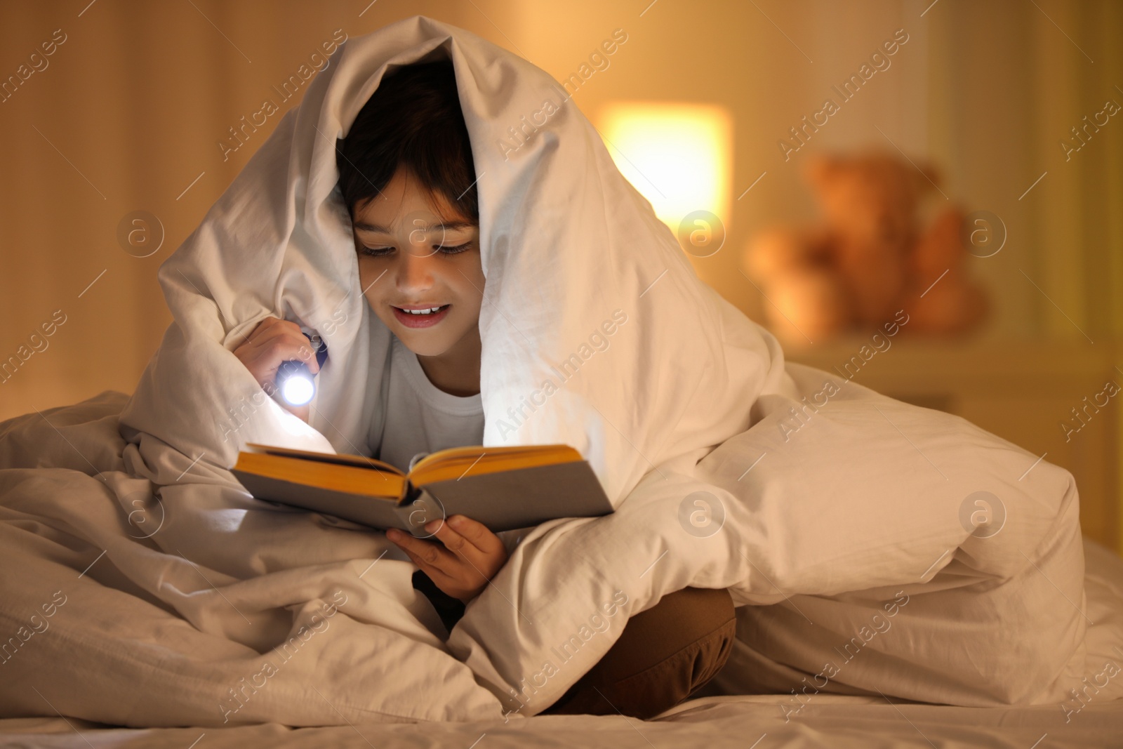 Photo of Boy with flashlight reading book under blanket at home