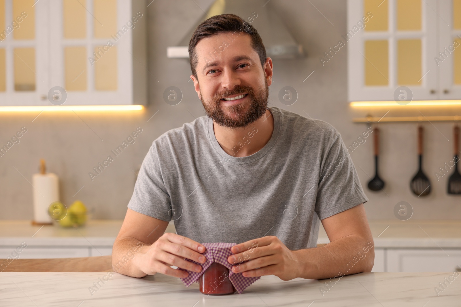 Photo of Man packing jar of jam into beeswax food wrap at table in kitchen