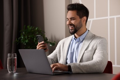 Happy young man with smartphone working on laptop at table in office