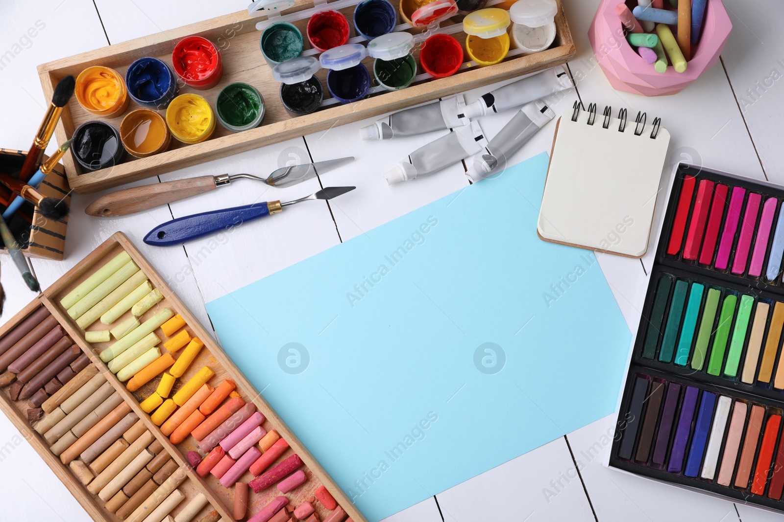 Photo of Blank sheet of paper, colorful chalk pastels and other drawing tools on white wooden table, flat lay. Modern artist's workplace