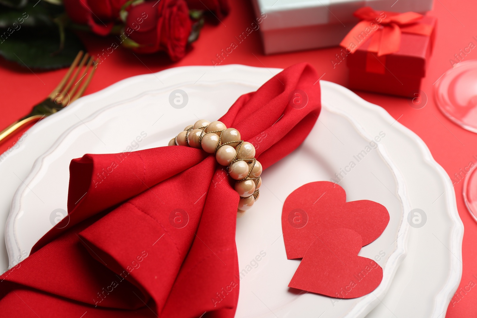 Photo of Plates with napkin, paper hearts, gift boxes and bouquet of roses on red table, closeup. Romantic dinner place setting