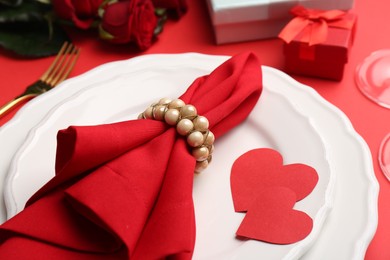 Photo of Plates with napkin, paper hearts, gift boxes and bouquet of roses on red table, closeup. Romantic dinner place setting