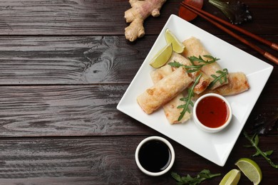 Photo of Tasty fried spring rolls, arugula, lime and sauces served on wooden table, flat lay. Space for text