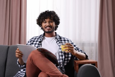 Photo of Happy man holding glass of juice and magazine on sofa with wooden armrest table at home