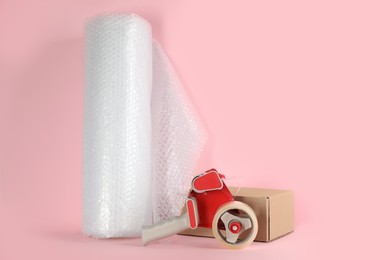 Photo of Bubble wrap roll, tape dispenser and cardboard box on pink background, space for text