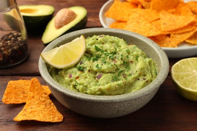 Photo of Bowl of delicious guacamole, lime and nachos chips on wooden table, closeup
