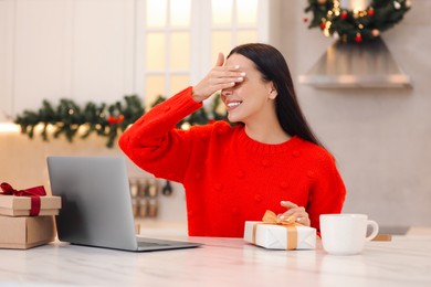 Photo of Celebrating Christmas online with exchanged by mail presents. Smiling woman covering eyes before opening gift box during video call at home