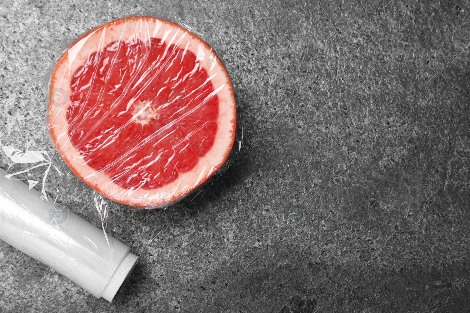 Photo of Half of grapefruit with plastic food wrap on grey table, flat lay. Space for text