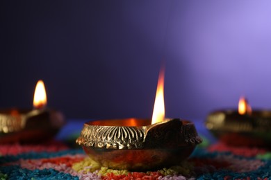 Photo of Diwali celebration. Diya lamps and colorful rangoli on table against violet background, closeup
