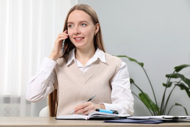 Photo of Woman taking notes while talking on smartphone at wooden table in office