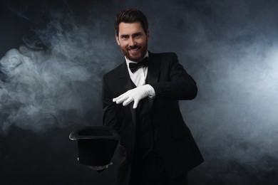 Photo of Happy magician showing magic trick with top hat in smoke on dark background