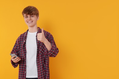 Teenage boy with smartphone showing thumb up on orange background. Space for text
