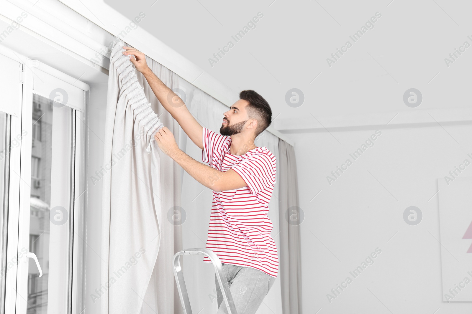 Photo of Young handsome man hanging window curtain in room