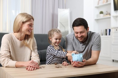 Family budget. Little girl putting coin into piggy bank while her parents watching indoors