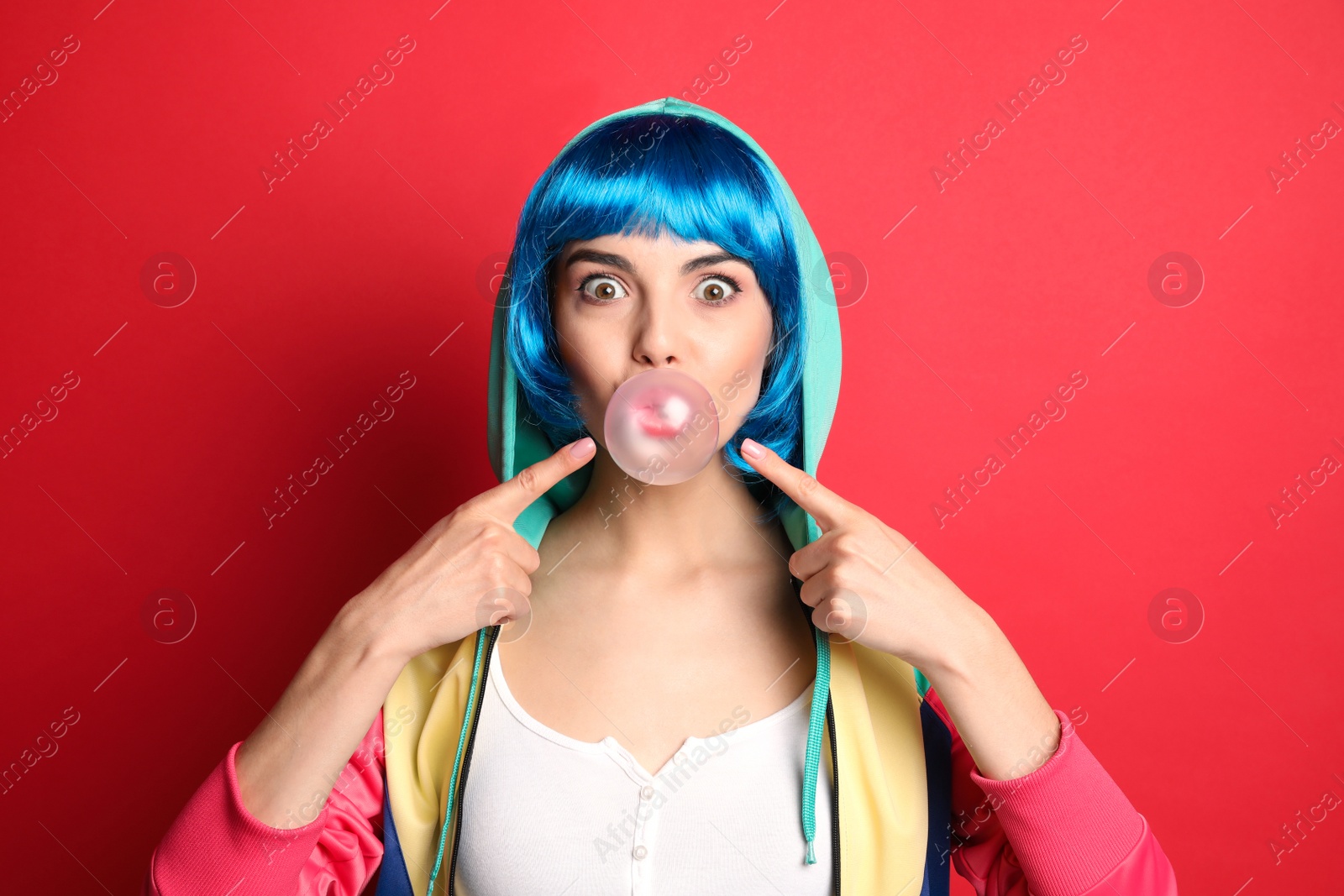 Photo of Fashionable young woman in colorful wig blowing bubblegum on red background