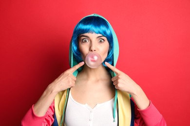 Photo of Fashionable young woman in colorful wig blowing bubblegum on red background