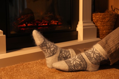 Woman in warm socks sitting near fireplace with burning wood at home, closeup