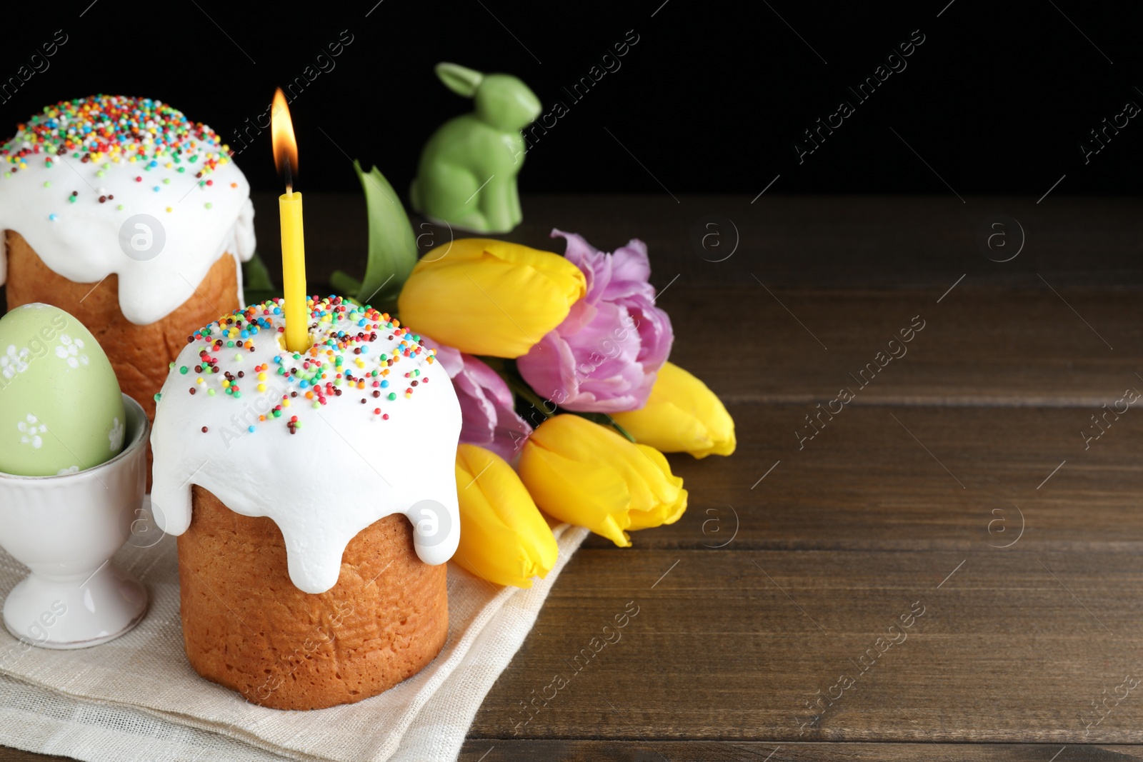 Photo of Traditional Easter cakes and one with burning candle on wooden table against black background. Space for text