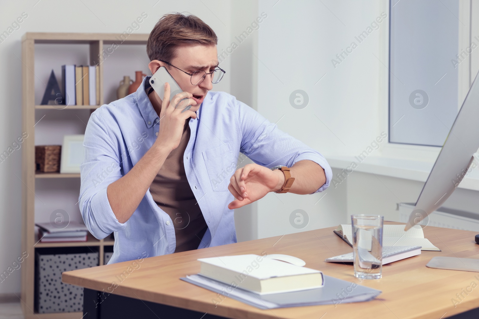 Photo of Emotional young man checking time while talking on phone in office. Being late